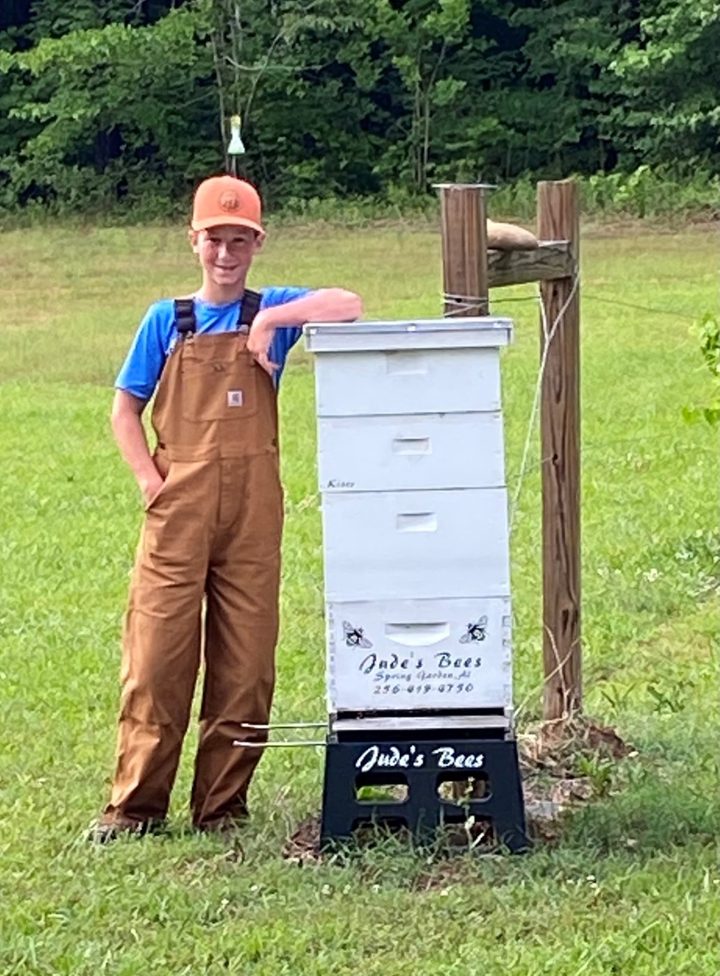 Jude in overalls, leaning on one of his Jude's Bees hives in the field